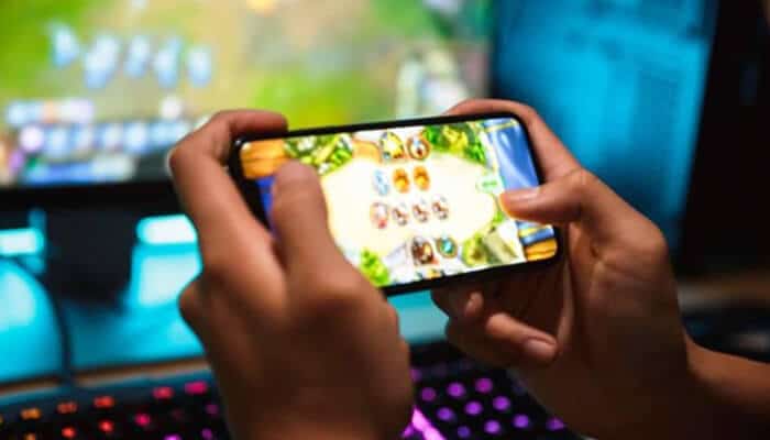 Online Slots Close-up of a person playing a mobile game with their hands, while a gaming monitor in the background suggests a gaming setup.