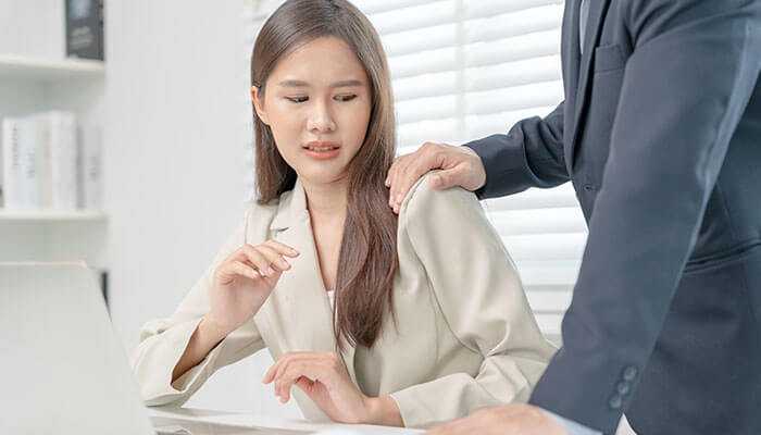 Women Uncomfortable woman at her desk looking at a man’s hand on her shoulder in an office setting.
