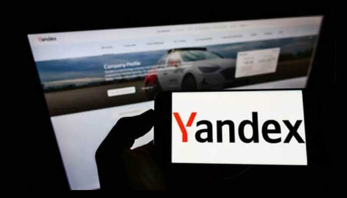 Yandex sells off russian business A hand holding a smartphone displaying the yandex logo, with a website company profile showing a self-driving car in the background.