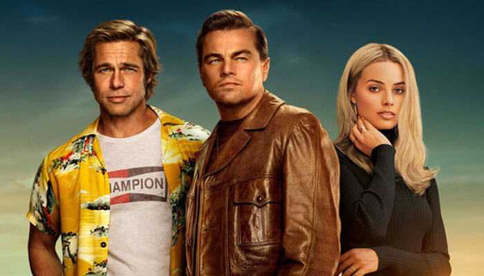 Once upon a time in hollywood - tycoonstory | tycoonstory media Three well-dressed individuals standing together against a sunset sky background: a man in a floral yellow shirt, a man in a brown leather jacket, and a woman with long blonde hair wearing a black outfit, all posing confidently side by side.