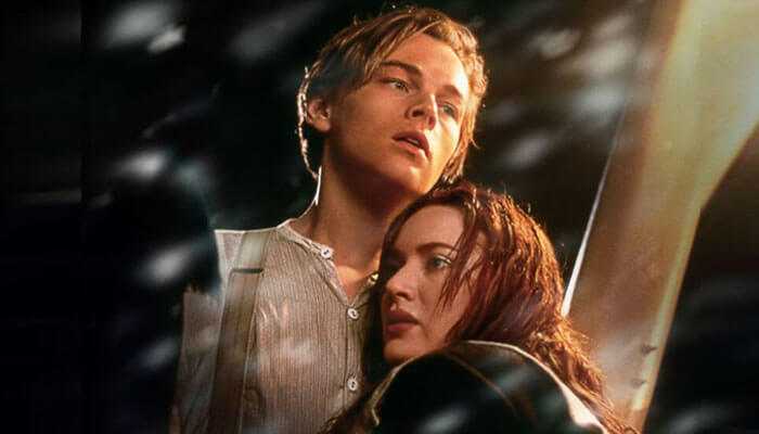 Leonardo dicaprio movies Two young adults holding onto each other on a ship’s deck in a dramatic, stormy scene, with light and shadows emphasizing the emotional moment.