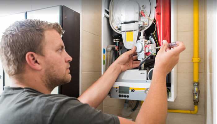 Check pilot light - tycoonstory | tycoonstory media Technician repairing a wall-mounted gas boiler, using a screwdriver to adjust internal components while inspecting the heating system.