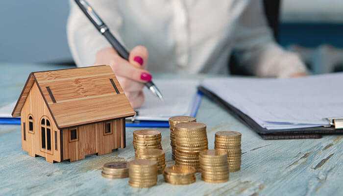 A close-up of a wooden house model beside stacks of coins, with a person writing notes in the background, symbolizing financial planning for buying a commercial property. Additional costs involved in buying a commercial property