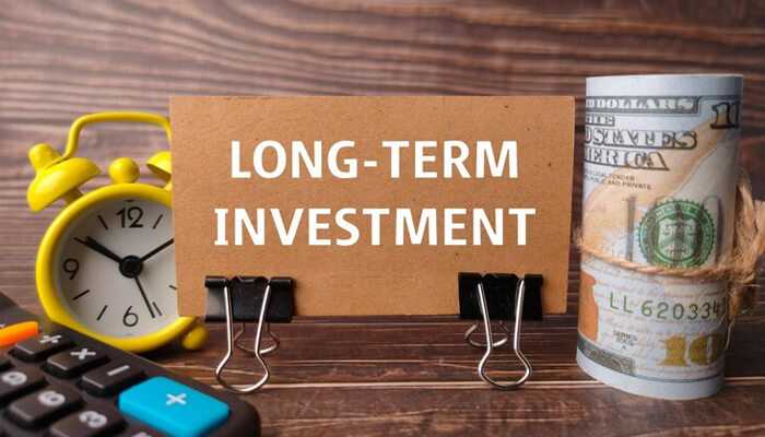 Adopt a long-term investment perspective - tycoonstory | tycoonstory media A table with a yellow color clock, a stack of us dollars and calculators, and cardboard labeled as a long-term investment symbolizes smart wealth management.