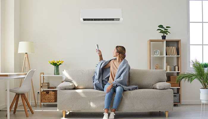 Energy efficiency and cost savings - tycoonstory | tycoonstory media Person sitting on a sofa using a remote to operate a split system air conditioner mounted on the wall illustrating home comfort and efficient cooling with split system air conditioning