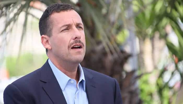 Adam sandler net worth 2024 - tycoonstory | tycoonstory media Adam sandler with short dark hair and a mustache, wearing a navy blazer and light blue shirt, standing outdoors with palm trees in the background