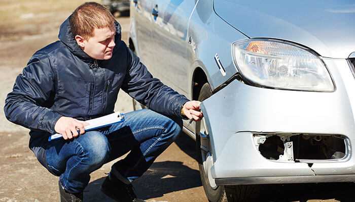 Collecting evidence - tycoonstory | tycoonstory media A man inspecting damage to the front bumper and side of a silver car after a car accident, holding a clipboard while crouching down, representing insurance work in a car accident