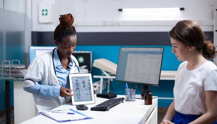Continuous support monitoring and patient empowerment - tycoonstory | tycoonstory media Wipro healthcare interoperability solutions: doctor consulting a patient, displaying an x-ray on a tablet in a modern clinic setting.