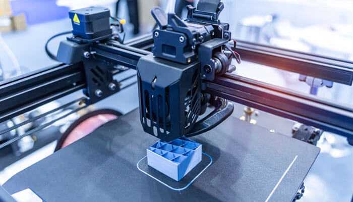 Cost-effective and sustainable production - tycoonstory | tycoonstory media Close-up of a 3d printer in light industry, building a small, blue and white grid-patterned object, demonstrating additive manufacturing.