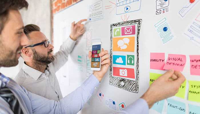 Mobile app development - tycoonstory | tycoonstory media Entrepreneurs brainstorming business ideas for entrepreneurs focused on mobile app design, using a whiteboard with sketches and notes to visualize user interface and functionality concepts