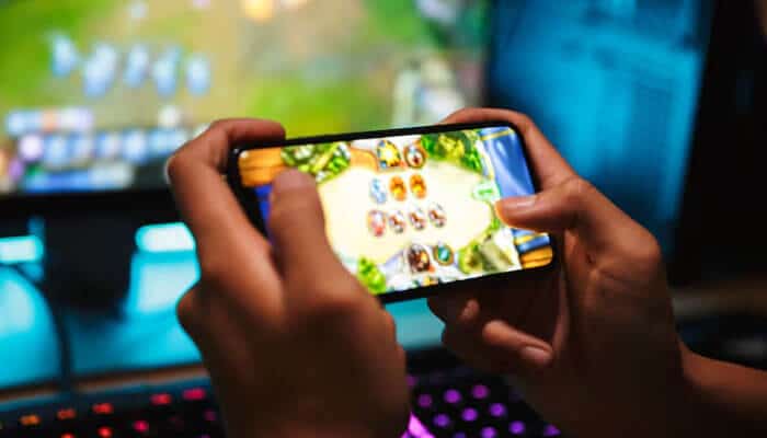 Mobile-friendly - tycoonstory | tycoonstory media Person playing a mobile game on a smartphone, showcasing the immersive experience of tiranga games with a gaming setup in the background
