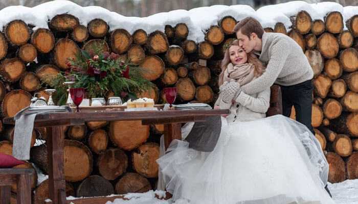 Rustic log cabin wedding - tycoonstory | tycoonstory media Wedding themes perfect for winter cozy outdoor snowy setting with a bride in a white gown groom in warm attire rustic wooden table red wine glasses and a log backdrop
