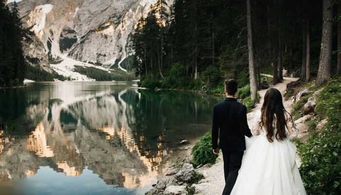 Snowy woodland wedding - tycoonstory | tycoonstory media Wedding themes perfect for winter romantic couple walking along a serene forest lake bride in a flowing white gown and groom in a classic black suit with snowy mountain reflections