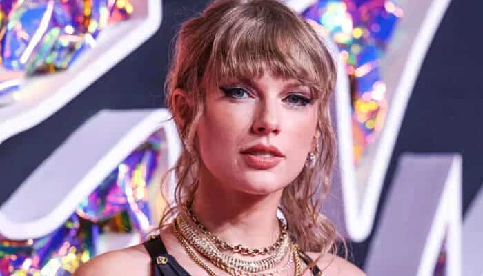 Taylor swift - tycoonstory | tycoonstory media Taylor swift with light-colored hair styled in loose bangs, wearing layered necklaces and a stylish outfit, posing against a vibrant, reflective backdrop. This is connected to the buzz surrounding taylor swift and travis kelce.