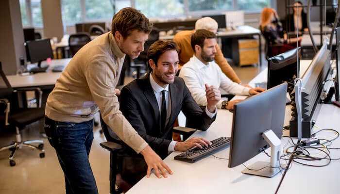 Verify the availability of their it experts - tycoonstory | tycoonstory media It firms for business support office team collaborating on a project, two men focus on a computer screen while discussing ideas in a modern open workspace with other team members working in the background