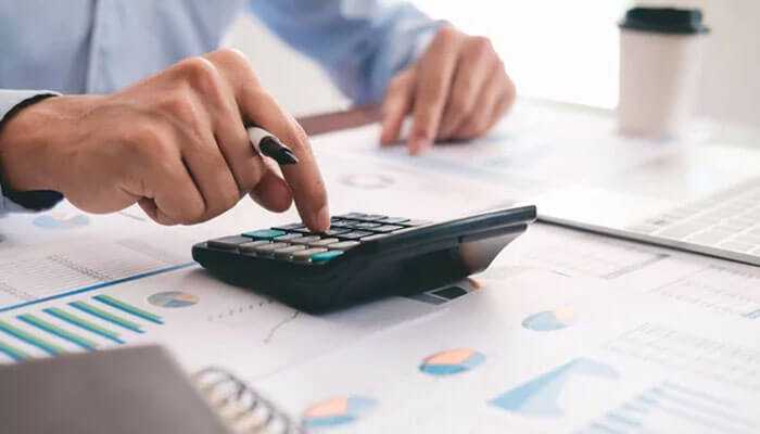 Cost savings - tycoonstory | tycoonstory media A person using a calculator on a desk with financial documents and charts, representing automating compliance reporting.