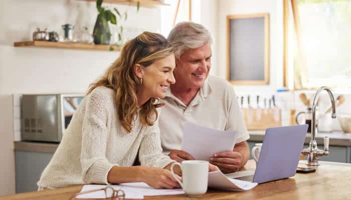 Creating multiple income streams - tycoonstory | tycoonstory media Smiling senior couple sitting at a kitchen table, reviewing documents and using a laptop, symbolizing retirement planning.