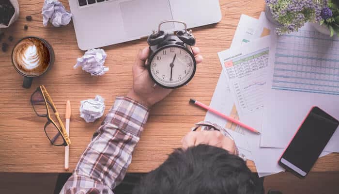 Discourage working off the clock - tycoonstory | tycoonstory media Digital wellness in the workplace a person holding an alarm clock at a cluttered desk, symbolizing time management and productivity