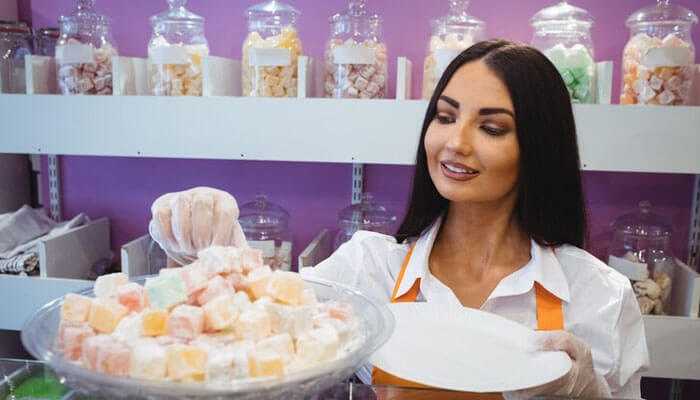 Exploring the impact of seasonal patterns - tycoonstory | tycoonstory media Shop worker selecting colorful candies from a display tray, illustrating the process of purchasing wholesale bulk sweets for retail sale.