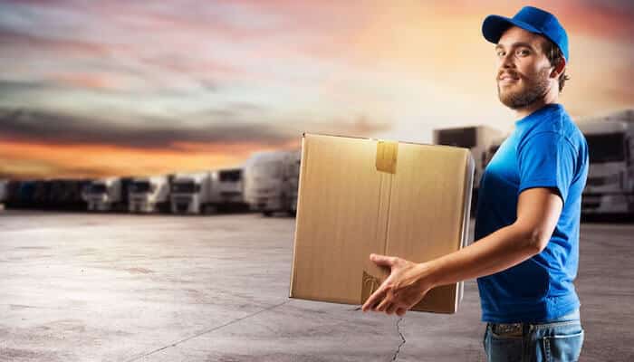 Facilitates the shipping of products - tycoonstory | tycoonstory media A delivery man in a blue uniform holding a cardboard box for product packaging, with trucks at sunset in the background.