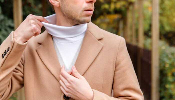 Layer over a collared shirt - tycoonstory | tycoonstory media Man adjusting a beige coat over a white turtleneck sweater, showing men's sweaters in an outdoor environment.