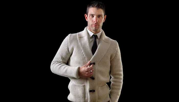 Layer with a blazer or suit jacket - tycoonstory | tycoonstory media Man in cream coloured buttoned cardigan over shirt and tie, posed against black background.