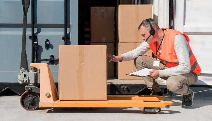 Logistical challenges - tycoonstory | tycoonstory media Shipping oversized goods a logistics worker wearing an orange safety vest and headset inspecting a large cardboard box on a manual pallet jack, with a clipboard in hand, standing near a truck dock