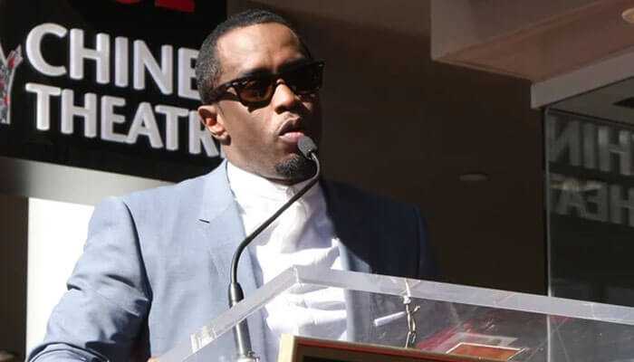 Media and entertainment ventures - tycoonstory | tycoonstory media The image depicts p diddy net worth wearing sunglasses and a light-colored suit speaking at a podium with a microphone. The background shows part of a sign reading chinese theatre