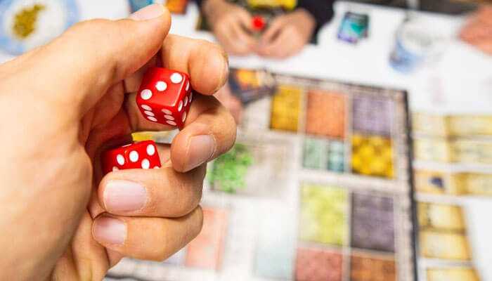 Must-play classic board games - tycoonstory | tycoonstory media Close-up of a hand holding two red dice with a blurred classic board games setup in the background, featuring colorful tiles and game pieces