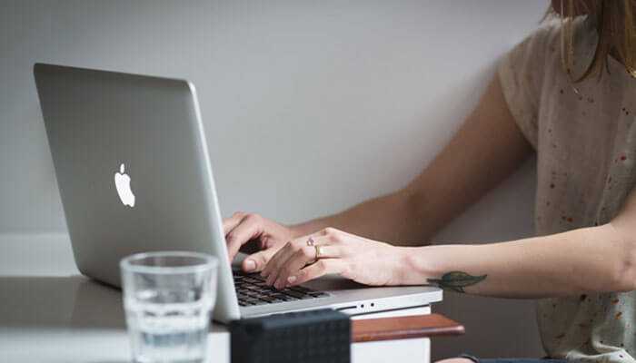 Offering free resources to showcase expertise - tycoonstory | tycoonstory media Person typing on a silver macbook at a desk with a glass of water, representing tasks common in service based businesses.