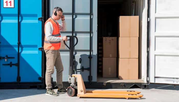Optimised packing - tycoonstory | tycoonstory media Worker wearing a high-visibility vest using a pallet jack to load boxes into shipping containers, illustrating efficient container loading practices