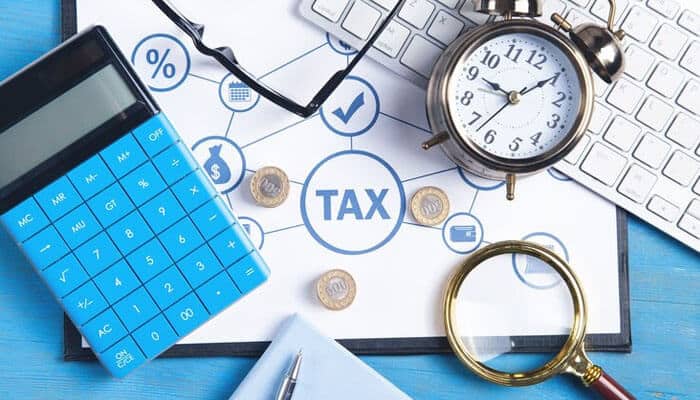 Tax benefits - tycoonstory | tycoonstory media Tax-related workspace with a calculator, clock, magnifying glass, keyboard, coins, and a diagram highlighting tax at the center, symbolizing financial, tax solutions, and term plan insurance