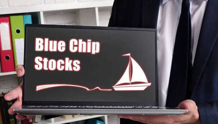 Top blue-chip stocks for conservative investors 2024 - tycoonstory | tycoonstory media A man in suit holding a laptop with blue chip stocks and a sailboat graphic on the screen.