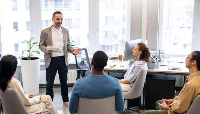 Train your team - tycoonstory | tycoonstory media A person leading a meeting or workshop in a modern office; colleagues are seated and engaged in the discussion.