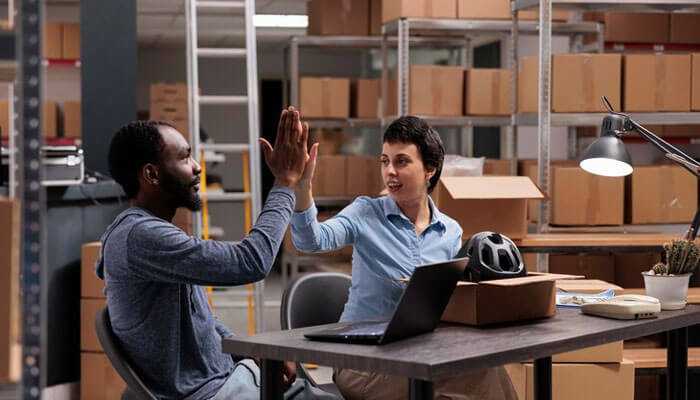 Training and support - tycoonstory | tycoonstory media Two high-fiving coworkers in a warehouse during the supplier onboarding process, with boxes and a laptop in the background.