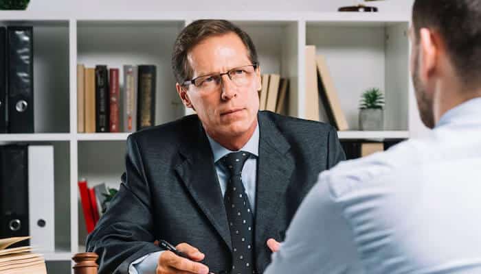 Consider their approach - tycoonstory | tycoonstory media A suit-wearing defense attorney listens attentively to a client in a meeting, with office bookshelves in the background.