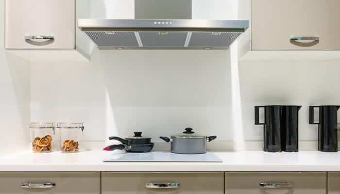 Cook without disturbance - tycoonstory | tycoonstory media A modern kitchen with a kitchen chimney, beige cabinets, a white countertop, jars, black kettles, and a pot and pan on the stove.