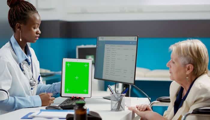 Improving financial and inventory management- tycoonstory | tycoonstory media Medspa software: a doctor holds a green-screen tablet while consulting with an older patient in a clinical setting, with a computer and office supplies visible