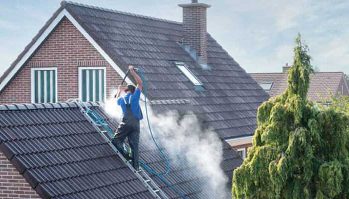 Roof cleaning and pressure washing - tycoonstory | tycoonstory media Dan ryan's Pressure Cleaning shows a worker pressure washing the tiled roof of a brick house while standing on a ladder, with mist from the cleaning visible and a large tree in the foreground.
