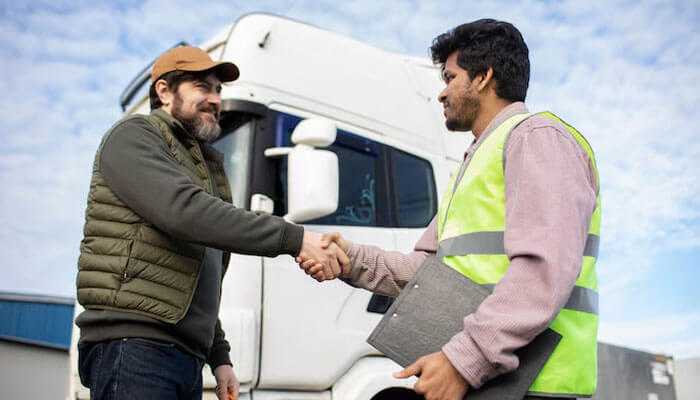 Stronger client relationships - tycoonstory | tycoonstory media A truck driver and a logistics worker shaking hands by a truck, symbolizing freight factoring for trucking success.