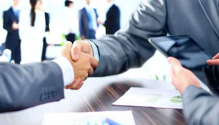 Support growth- tycoonstory | tycoonstory media Two businessmen from small businesses in singapore shaking hands over a table with papers and a tablet to seal an agreement in the corporate world.
