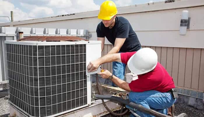 The benefits of hvac training for immigrants | tycoonstory media Two workers on a rooftop repair an hvac unit, demonstrating skills from hvac training programs. One wears a yellow hard hat, the other a white hard hat. It's a sunny day.