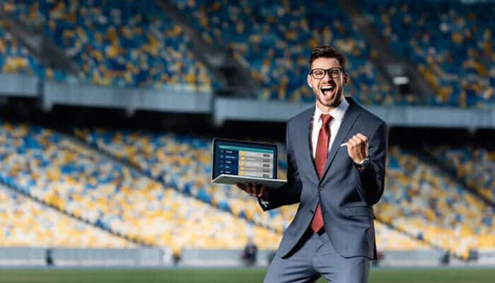 There is a prospect of winning - tycoonstory | tycoonstory media Excited tech-savvy gamer in india, in a suit, holding a laptop displaying sports betting odds in a stadium, celebrating a successful bet.