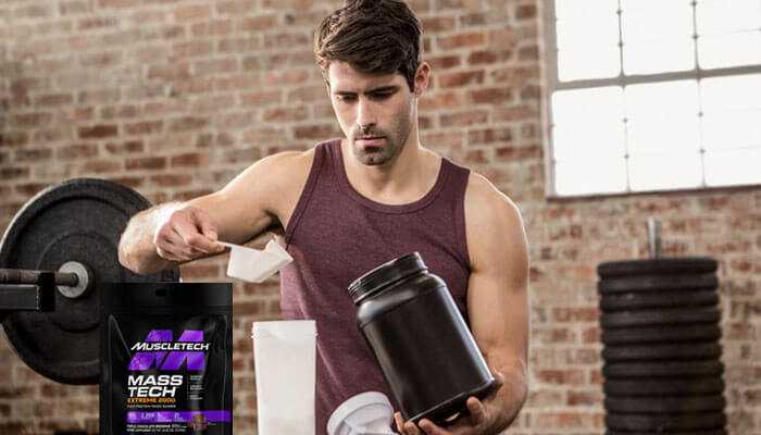 Time your mass gainer intake - tycoonstory | tycoonstory media A man in the gym with a maroon tank top on, shaking a protein shake. Mass gainers muscletech mass tech extreme 2000.