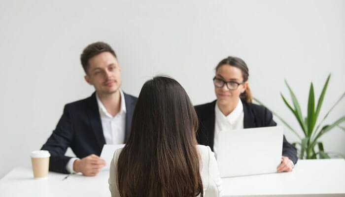 Training and onboarding new reps - tycoonstory | tycoonstory media A woman sits facing two interviewers discussing common sales challenges in a professional office setting with a plant.
