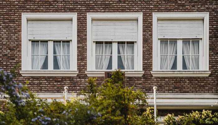 Assortment of styles to choose from - tycoonstory 1 | tycoonstory media A brick building with three windows, partially closed white roller shutters, white curtains, and outdoor blinds. It is surrounded by greenery.
