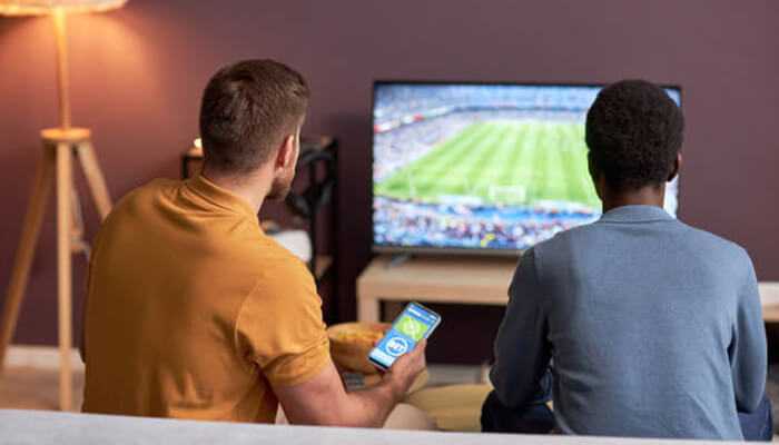 Live streaming and in-play betting- tycoonstory | tycoonstory media Two men watching a live football match on tv while one checks a sports betting app on his smartphone, illustrating the rise of live streaming and in-play betting in india’s sports betting industry