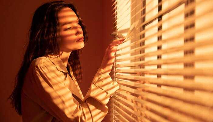 Timber venetians - tycoonstory | tycoonstory media A woman standing near a window, gently adjusting the blinds as warm sunlight filters through, showcasing various window blind options for a cozy and stylish ambiance.