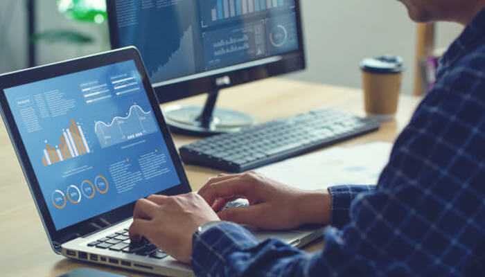 V | tycoonstory media A person in a blue checkered shirt analyzing financial charts on a laptop, with a desktop monitor displaying market data in the background.