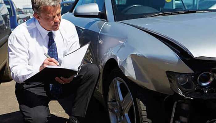 Delaying the insurance claim - tycoonstory | tycoonstory media An insurance adjuster inspects and takes notes on a damaged silver car after a hit and run accident, with visible dents and a broken headlight.
